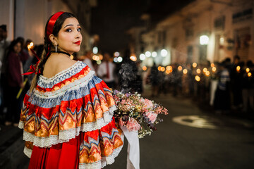Mujer sahumadora vestida con traje típico tradicional participa en una procesión nocturna durante la Semana Santa en Popayán, Colombia. Sostiene un ramo de flores mientras esparce sahumerio 