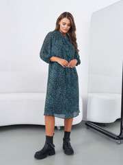 A woman wearing a dark patterned dress and black boots stands indoors, looking down. Her long hair is loose, and she's near a full-length mirror with a white couch behind her