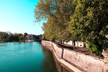 Fototapeta premium Serene Waterway with Lush Trees and Blossoming Foliage Along the Bank, Inviting a Peaceful Stroll on the Walkway, Leading to a Charming House with a Scenic View of the Calm Water and Vibrant Nature 