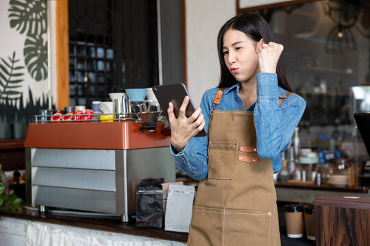 Asian young female barista wearing apron and denim shirt standing in coffee shop holding tablet with raised fist smiling proudly showing success motivation and positive energy in small business