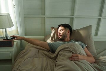 Fototapeta premium Asian man lying in bed under blanket reaching to turn off smartphone alarm with sleepy face in early morning bedroom with soft window light and calm peaceful atmosphere.