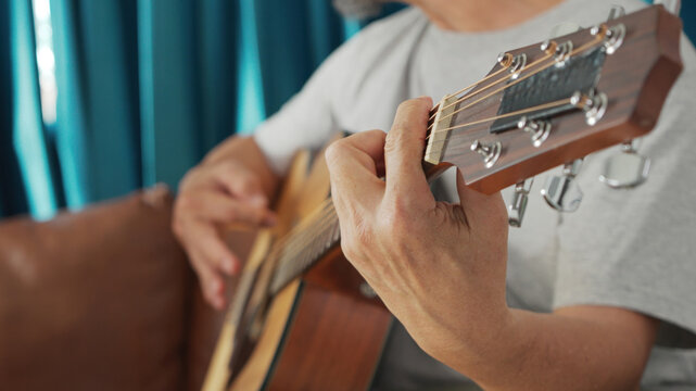 Close up Asian Senior man hands playing acoustic guitar, showcasing skillful finger placement while sitting on a sofa.