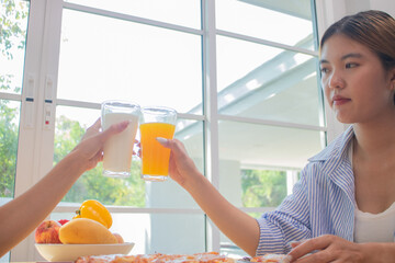 Two elegant young women sit in a house, smiling and clinking glasses of juice and breakfast of...