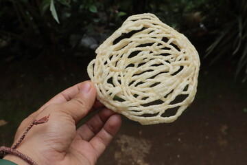 Hand holding crispy noodlelike snack with tangled strands set against an outdoor backdrop