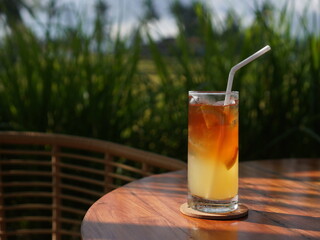 Iced tea with lemon and ice cubes served in a tall glass with a straw, placed on a wooden table outside. The green background adds a natural and serene ambiance.