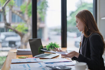 Businesswoman reviews paperwork and laptop on cluttered office desk now.