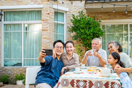 Happy Asian three generation family using mobile phone taking selfie together in the garden. Big family enjoy and fun celebration holiday event afternoon tea party and spending time together at home.