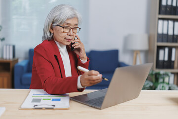 Focused asian elderly businesswoman in red suit talking on smartphone and pointing at laptop with pen while working with documents at wooden table in modern office