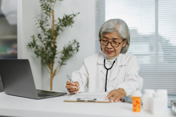 Senior female doctor wearing lab coat and stethoscope compiling medical records using laptop and taking notes on clipboard in her bright and modern office