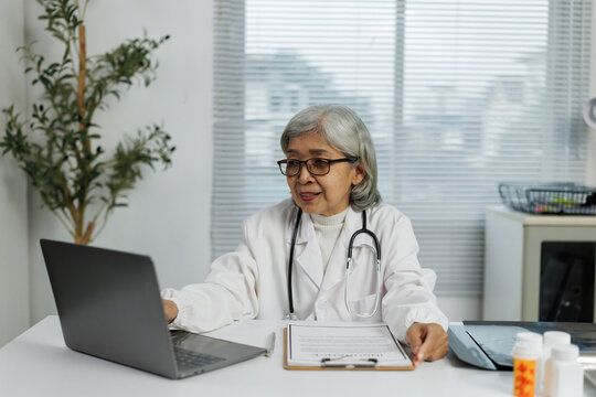 Asian female doctor wearing lab coat and stethoscope sitting at desk using laptop computer with medical record, x-ray image and medicine on table in hospital clinic office - Powered by Adobe