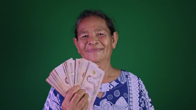 Happy elderly Asian woman holding Thai baht banknotes with big smile, Concept of financial success, luck, and retirement joy on chroma key green screen background, Grandmother happily with a money