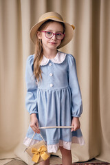 Stylish young girl in blue dress with long sleeves and glasses holds wicker handbag decorated with yellow bow. Model in straw hat smiles sweetly on fabric background.