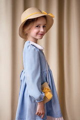A cute child in a straw hat with a yellow bow looks at the camera and smiles. The girl wears a blue dress and holds a small wicker shoulder bag decorated with a yellow bow. The background is neutral