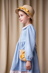 A sweet young girl in a romantic dress stands sideways and looks to the left. On her head she has a straw hat with a yellow ribbon and bow. Shot from the bottom up against a plain light curtain.