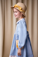 A funny little girl in profile on a fabric background. The child is looking to the left and smiling in a cheerful mood. The model is wearing a dress and a hat with a ribbon.