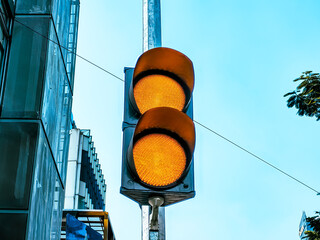 Close-up of a yellow traffic light glowing on a city street, signaling caution to drivers amid urban buildings and clear blue sky.