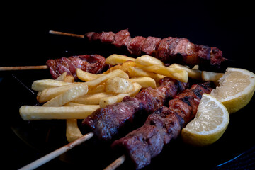 Greek souvlaki pork skewers with fried potatoes and slices of lemon inside a black plate.