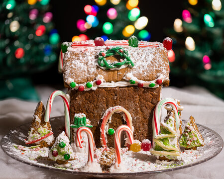 Homemade gingerbread house decorated with candy canes, powdered sugar and jelly beans with christmas trees blurred int he background