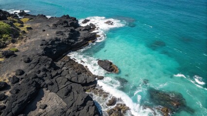 Obraz premium Top-down view of black volcanic rocks surrounded by bright turquoise ocean with foamy waves crashing along rugged coast. 