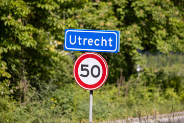 A clear "Utrecht" city entrance sign with a 50 km/h speed limit, set against a backdrop of vibrant green foliage under a sunny sky.