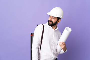 Young architect man with helmet and holding blueprints over isolated purple background . Portrait