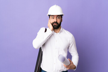 Young architect man with helmet and holding blueprints over isolated purple background frustrated...