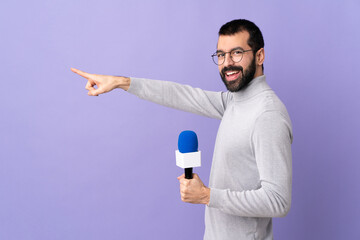 Adult reporter man with beard holding a microphone over isolated purple background pointing finger...