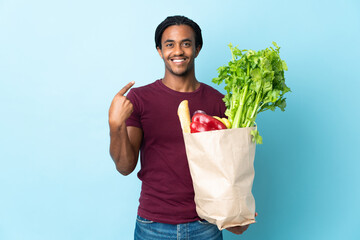 African American man holding a grocery shopping bag isolated on blue background with surprise...