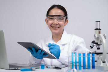 A smiling scientist in a lab coat uses a tablet, surrounded by test tubes and a microscope, showcasing modern research and technology in a laboratory setting.