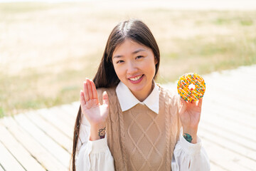Young Chinese woman holding a donut at outdoors saluting with hand with happy expression
