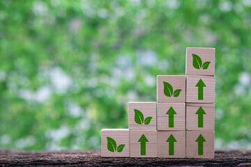 Stacked wooden blocks with green leaf symbols and arrows, representing growth and sustainability against a blurred green background.