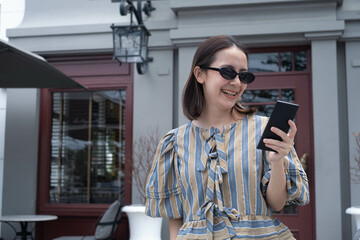 A cheerful young woman in stylish attire smiles while looking at her smartphone outside a cozy cafe.