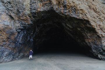Man exploring a large dark cave entrance on a sandy beach, documenting the natural wonder with his...