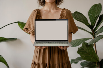 Woman in Earthy-Toned Dress Presenting Laptop with Blank Screen, Cozy and Natural Setting