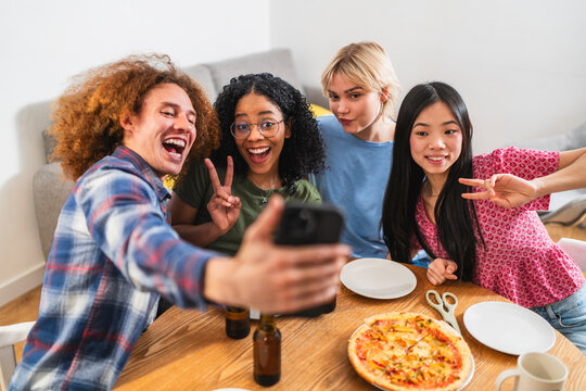 Four cheerful young multi ethnic friends are taking a selfie with a smartphone while enjoying pizza and drinks at home