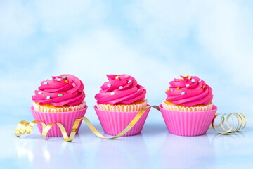 Pink frosted cupcakes with colorful sprinkles and golden ribbon on light blue background.