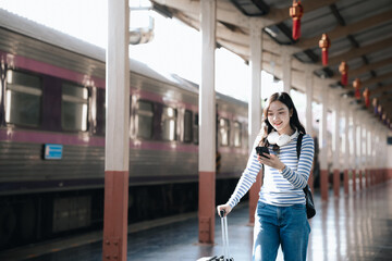 Fototapeta premium Asian woman sitting at a railroad station with backpack, using mobile phone and laptop, waiting for train, enjoying travel lifestyle.