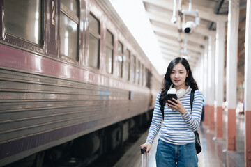 Asian woman sitting at a railroad station with backpack, using mobile phone and laptop, waiting for train, enjoying travel lifestyle.