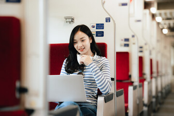 Obraz premium Asian woman sitting at a railroad station with backpack, using mobile phone and laptop, waiting for train, enjoying travel lifestyle.