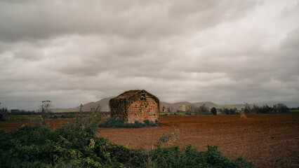 An abandoned stone house in the middle of a plowed field, under a dramatic cloudy sky. In the background you can see soft hills and country vegetation. Silent and melancholic atmosphere.