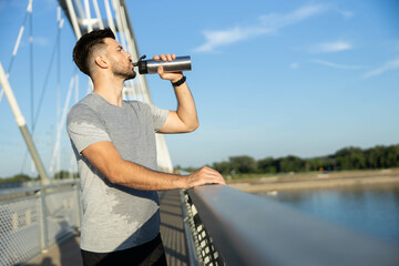 A man stands on a bridge, taking a refreshing sip from his water bottle while enjoying the view and...