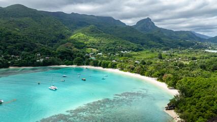 A green hillside overlooks a tranquil turquoise bay, with boats dotting the clear water. Butzel Beach. Seychelles, Mahe.