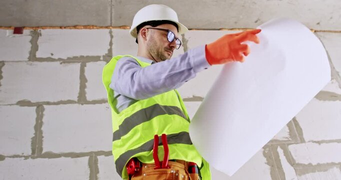 Man constructor inspects ceiling and building scheme before renovation. Specialist in helmet holds floor plan in hands assessing work scope against slag block wall