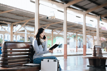 Fototapeta premium Asian woman sitting at a railroad station with backpack, using mobile phone and laptop, waiting for train, enjoying travel lifestyle.