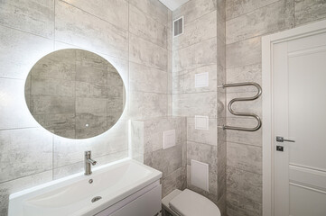 Modern bathroom featuring gray tiles, a round LED mirror, white sink, toilet, and door, plus a...