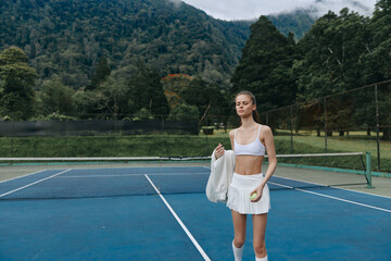 Young female tennis player in white athletic wear on a blue tennis court with a scenic mountain...