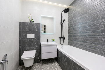 Modern bathroom with gray and white tiles, a floating vanity with a lit mirror, toilet, and a tub with a black shower fixture