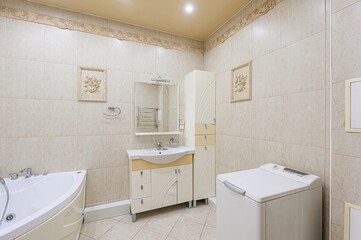 bright bathroom features a corner bathtub, a vanity with mirror, and a tall cabinet. The walls are tiled in a neutral color