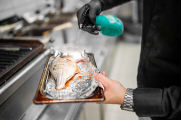 A professional chef seasons a fresh fish on a baking tray covered with aluminum foil in a commercial kitchen, showcasing culinary skills and food preparation techniques.