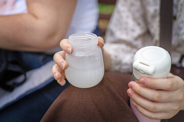 Young mother cooling baby bottle with hot water from a thermos in a park, preparing infant formula safely outdoors